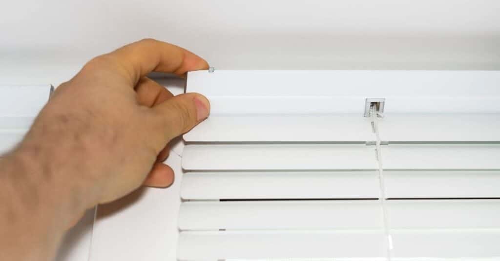 a left hand of a man shows about to remove a window blind, as the result of a homeonwer's search for "blind cleaning service near me"