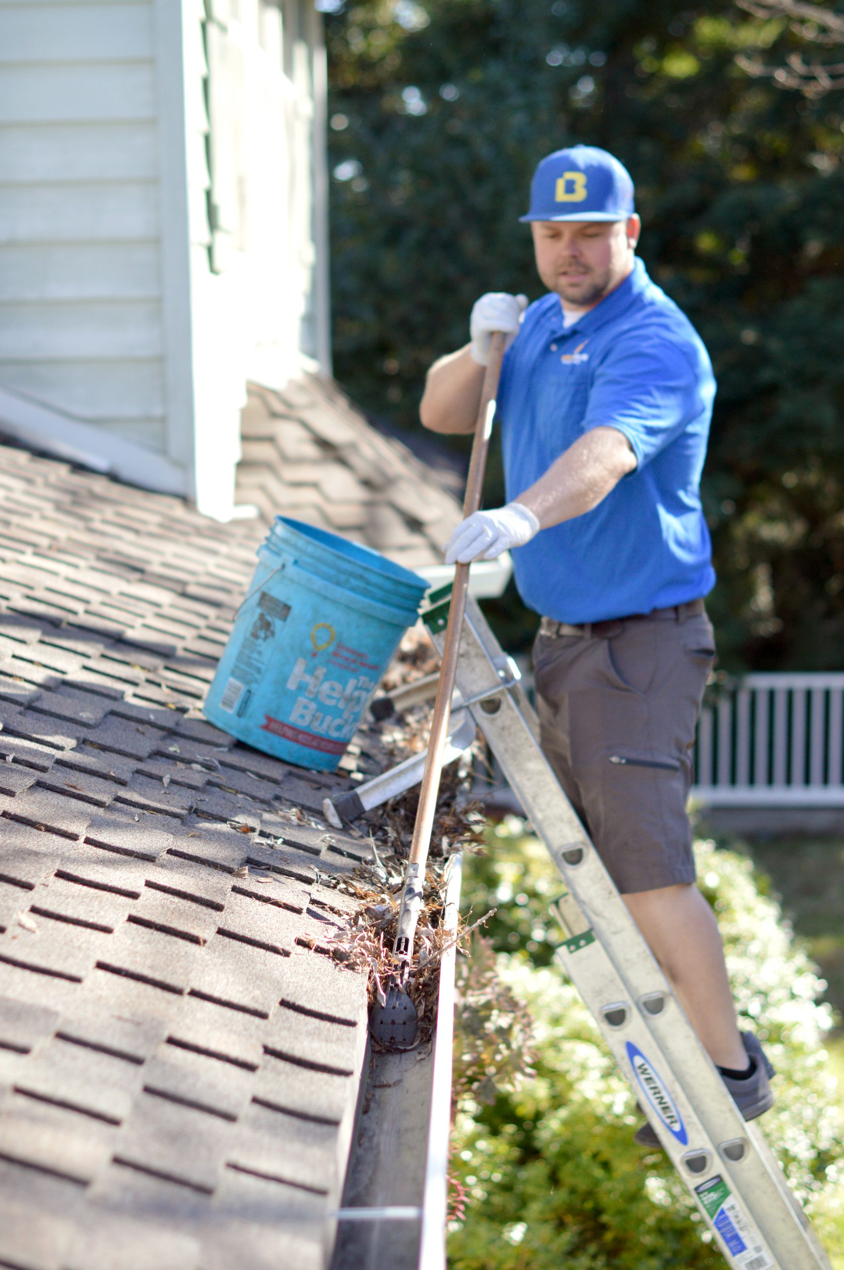 A Bailey Boys technician scooping leaves out of a roofline gutter while standing on a ladder